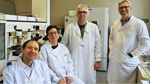 Three Men And A Woman Standing In A Laboratory All Wearing White Lab Coats. They Are Smiling